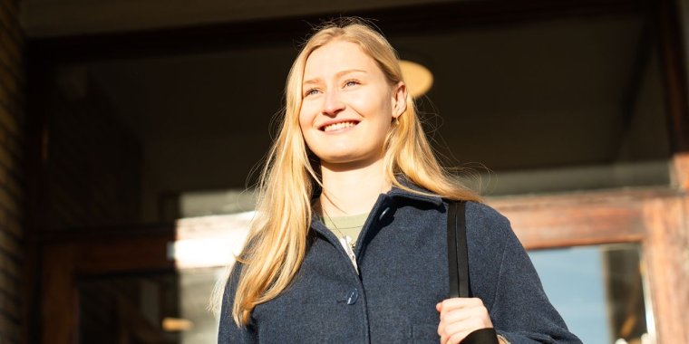 Kvinne med langt blondt hår smiler i solskinnet, iført blå jakke og bærer en veske.