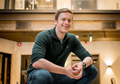 Mann smiler og sitter på en stol i et moderne innendørs miljø.