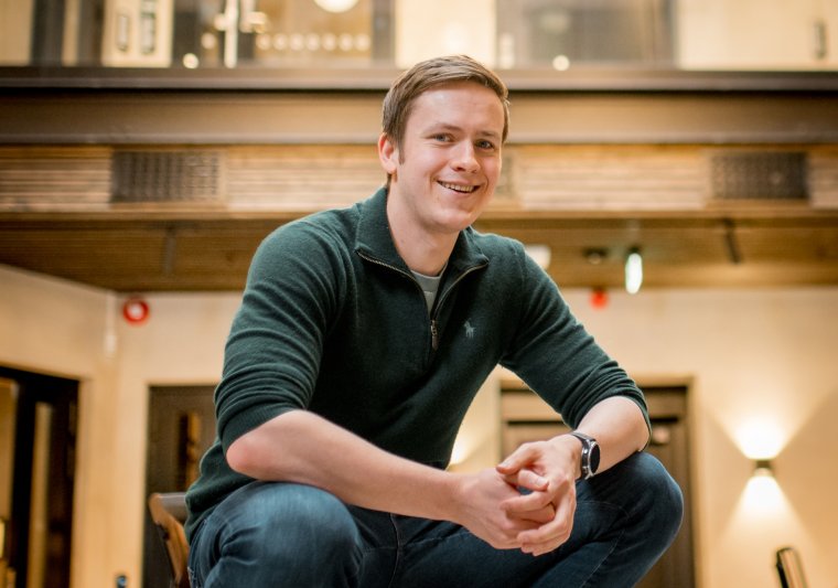 Mann smiler og sitter på en stol i et moderne innendørs miljø.