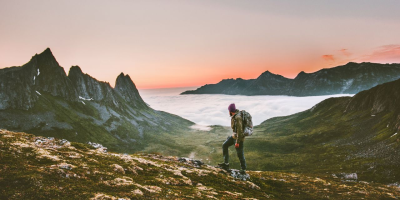 Person på fjelltur ved solnedgang med fjell og skyer i bakgrunnen.