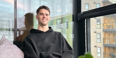 Mann i svart hettegenser smiler mens han sitter i en sofa ved et vindu.