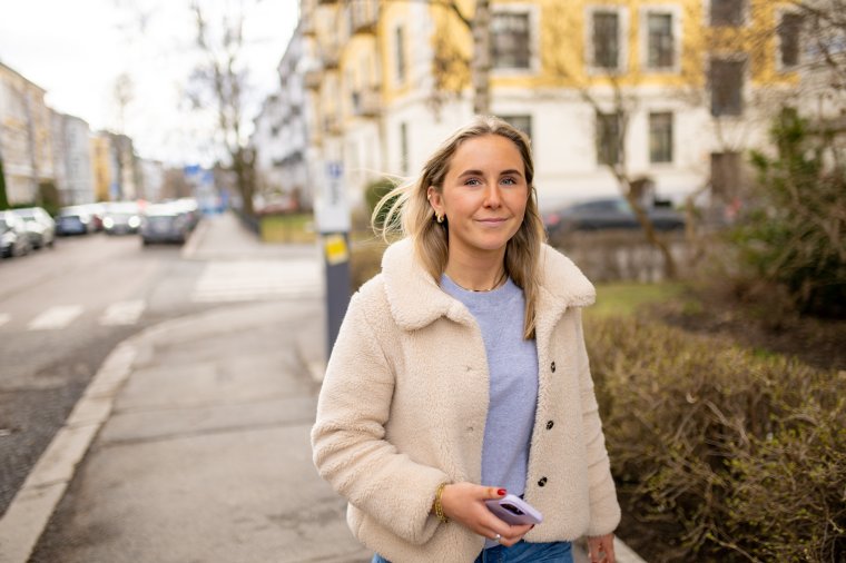 Kvinne i lys jakke går langs en gate med bygninger i bakgrunnen.