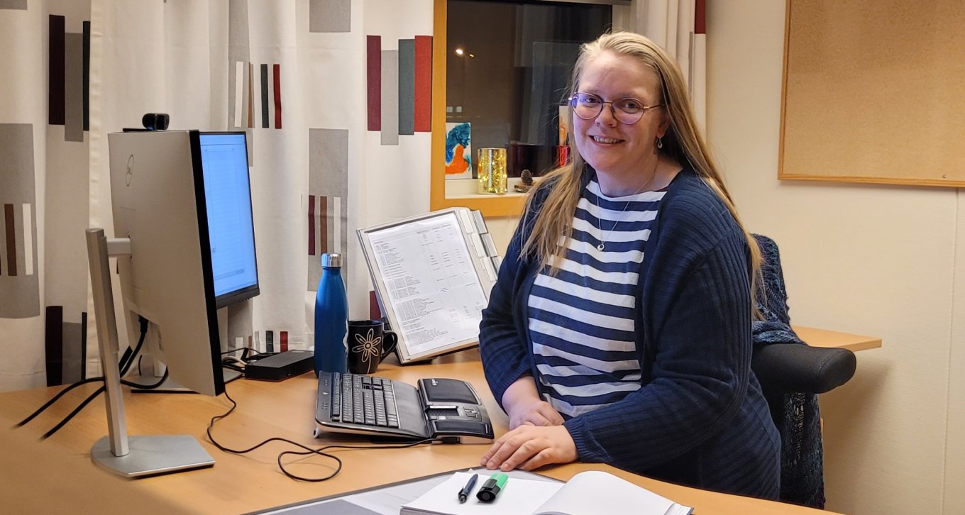 Kvinne smiler ved skrivebord med datamaskin, notatbok og vannflaske.