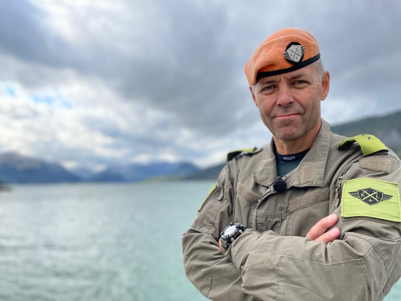 Mann i militæruniform med oransje beret foran en innsjø og fjell i bakgrunnen.