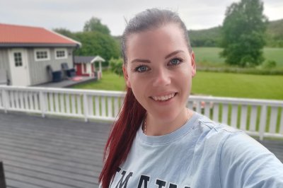 Kvinne smiler på veranda med grønn hage og hus i bakgrunnen.