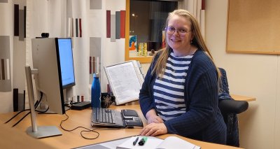 Kvinne smiler ved skrivebord med datamaskin, notatbok og vannflaske.