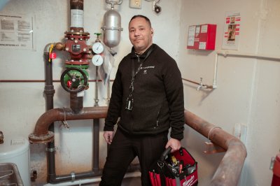 Person i teknisk rom med verktøyveske, rør og måleinstrumenter i bakgrunnen.