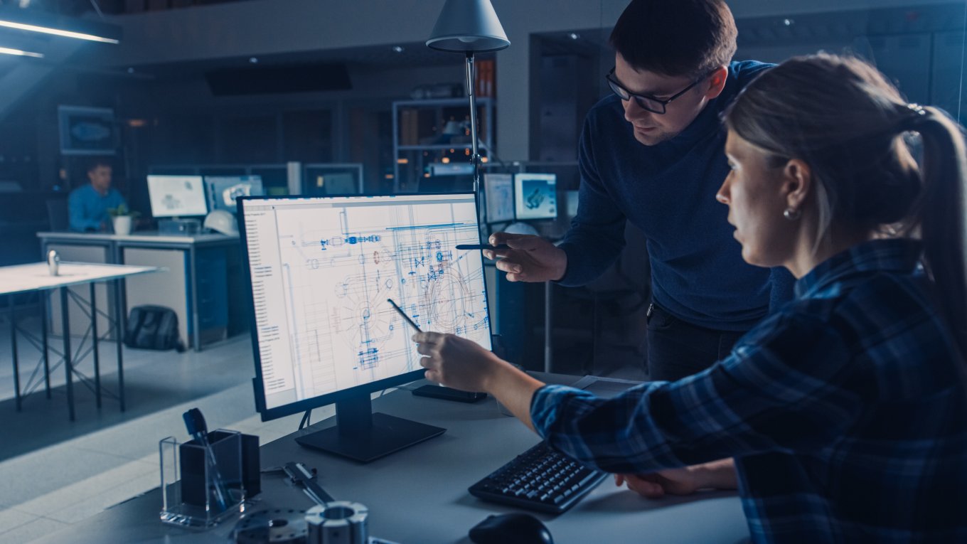 To personer diskuterer ingeniørtegninger på en dataskjerm i et moderne kontor.