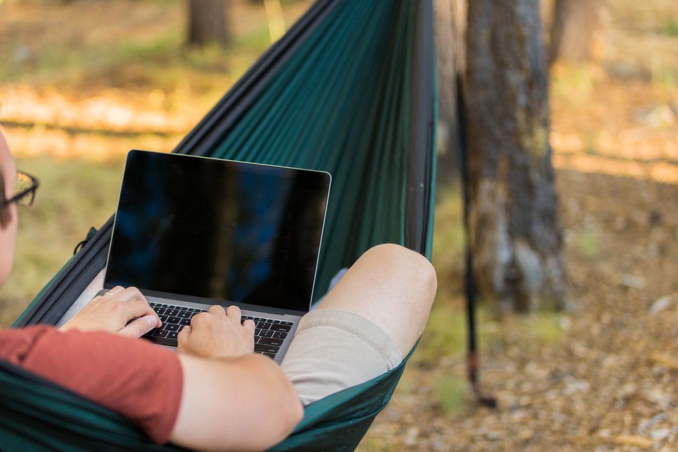 En mann jobber på PC i hengekøye ute i skogen