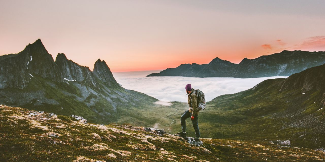 Person på fjelltur ved solnedgang med fjell og skyer i bakgrunnen.