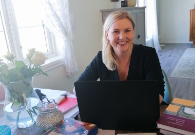 Kvinne smiler foran en bærbar PC med bøker og notater på bordet.