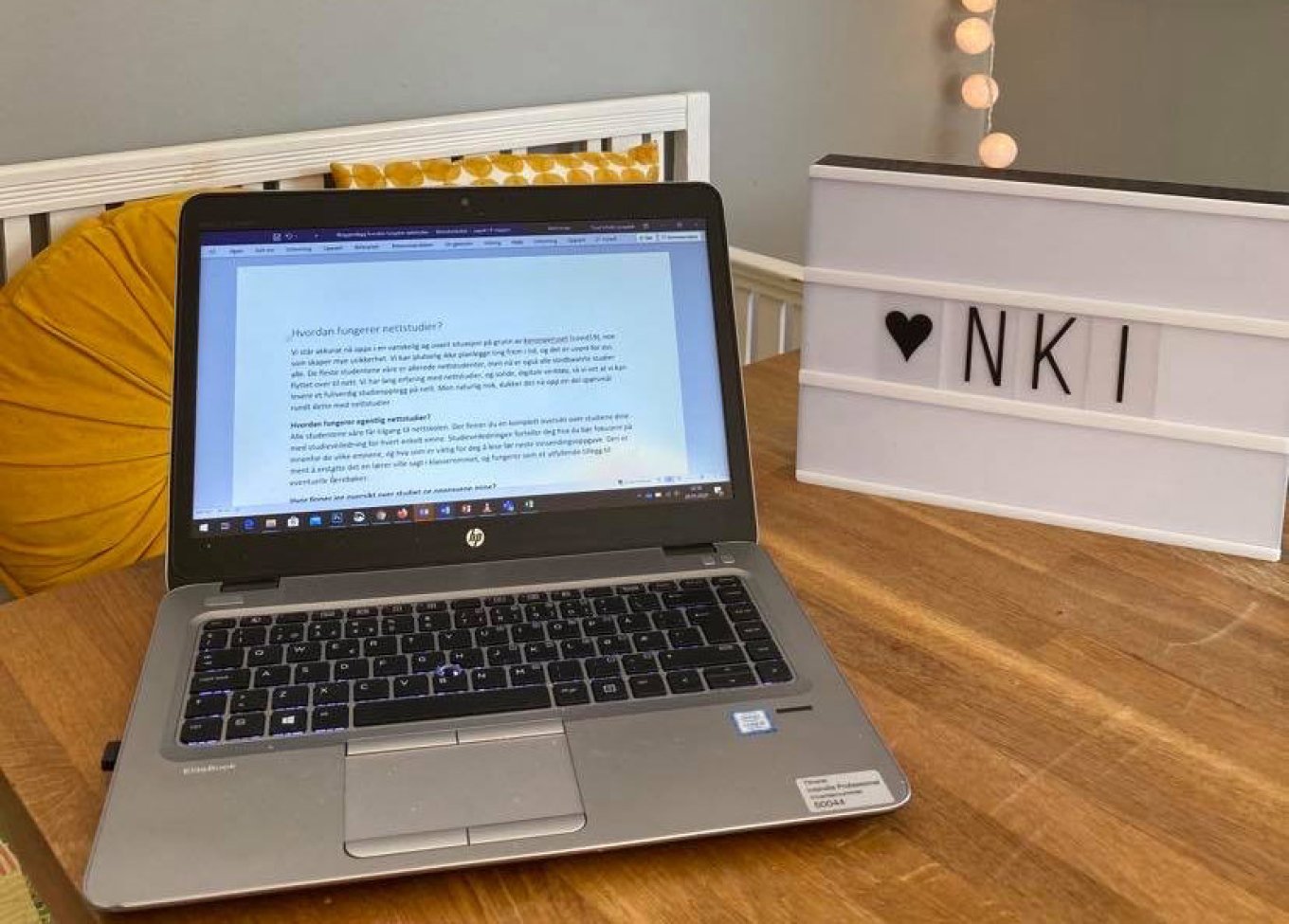 Bærbar PC på skrivebord med NKI-skilt i bakgrunnen.