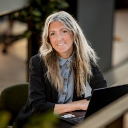 Kvinne med langt hår smiler mens hun jobber på en bærbar datamaskin.