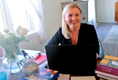 Kvinne smiler foran en bærbar datamaskin med bøker og blomster på bordet.