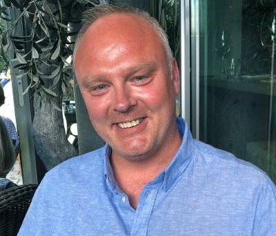 Mann i blå skjorte smiler foran et tre og et vindu.
