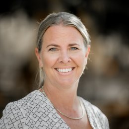 Kvinne med blondt hår og mønstret bluse smiler foran en uskarp bakgrunn.