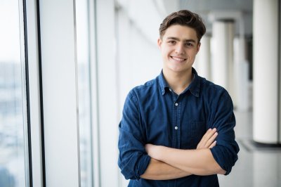 Ung mann i blå skjorte smiler og står med armene i kors ved et vindu.