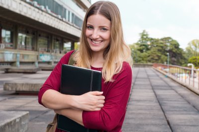 Smilende kvinne med bok foran bygning, klar for studier.