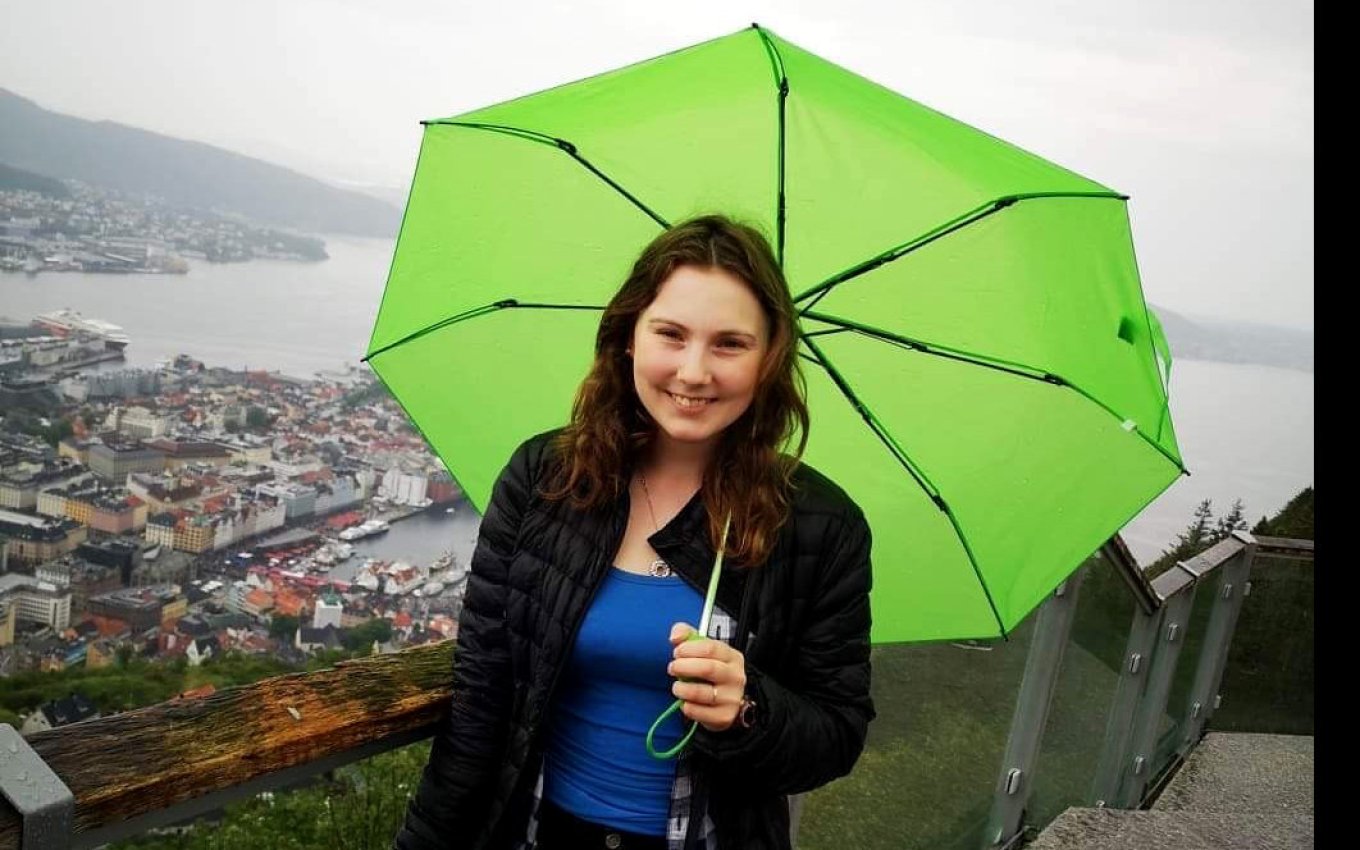 Kvinne med grønn paraply smiler foran en byutsikt på en overskyet dag.