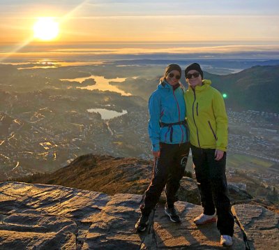 To personer på fjelltur med solnedgang og utsikt over by og fjord.