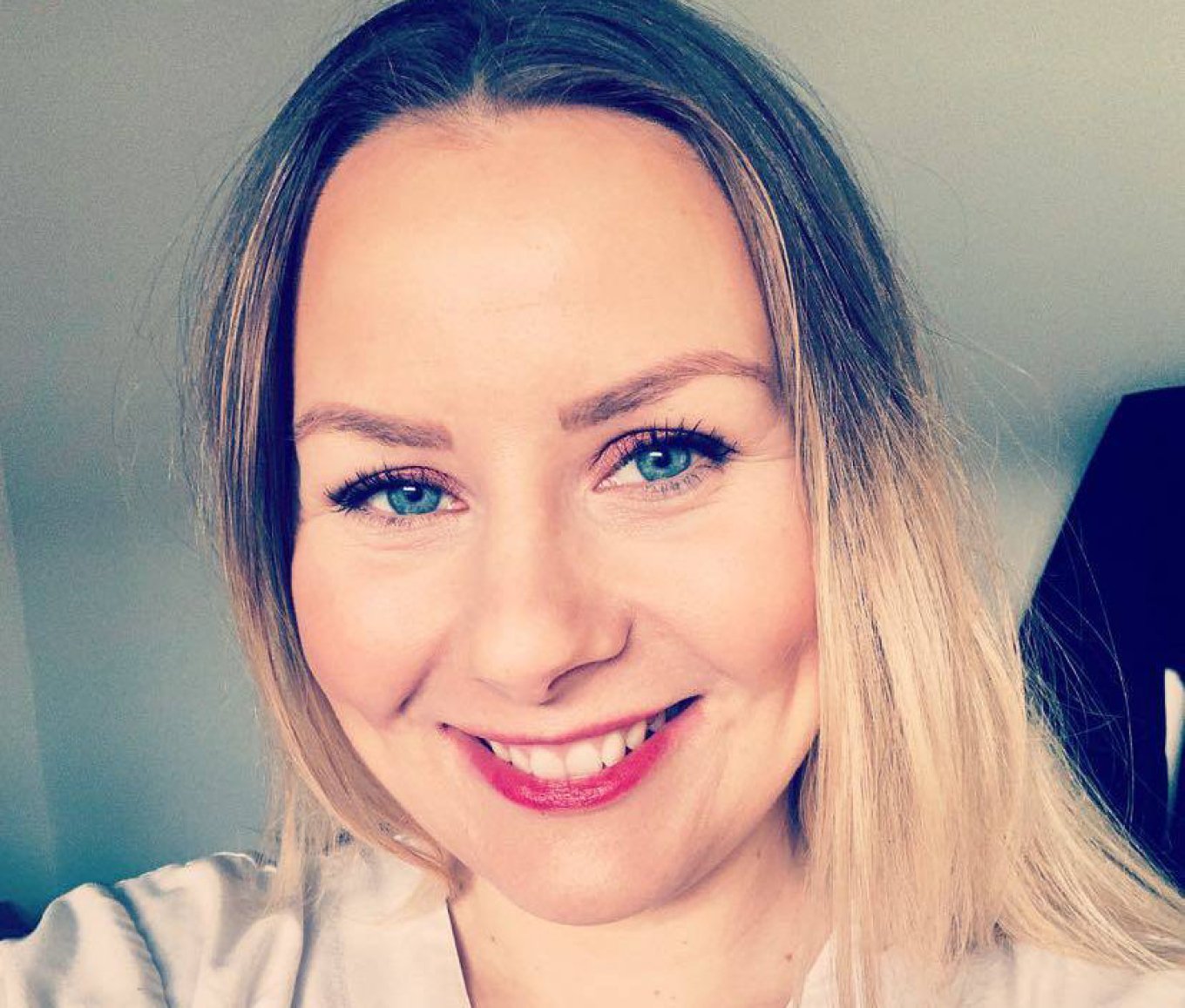 Kvinne med langt blondt hår smiler mot kameraet i et godt opplyst rom.