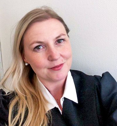 Kvinne med langt blondt hår og sort kappe smiler foran en hvit vegg.
