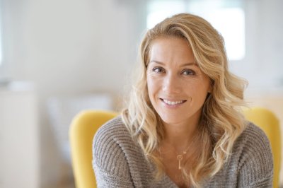 Kvinne med blondt hår smiler og sitter i en gul stol i et lyst rom.