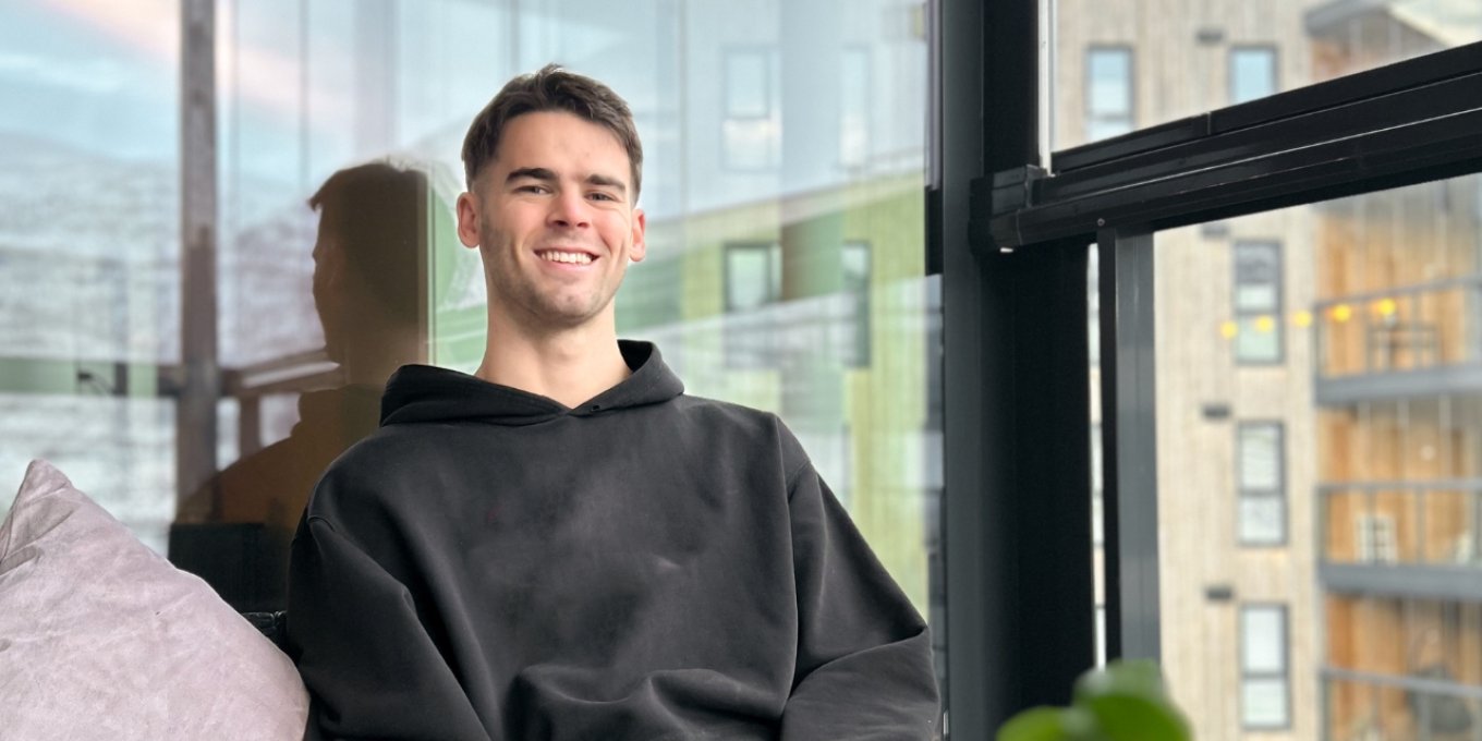Mann i svart hettegenser smiler mens han sitter i en sofa ved et vindu.