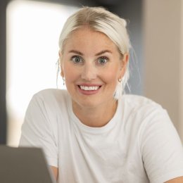 Kvinne med hvit t-skjorte smiler foran en laptop.