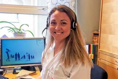Kvinne i hvit uniform med headset smiler foran datamaskin på kontor.