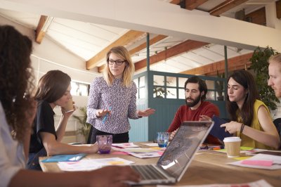 En gruppe studenter diskuterer rundt et bord med en kvinnelig leder som står og forklarer.