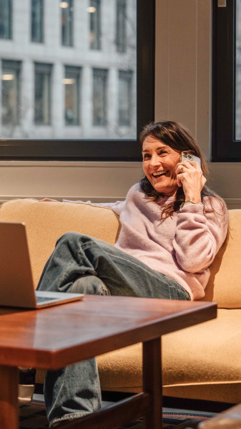 Kvinne smiler og snakker i telefonen mens hun sitter på en sofa med en laptop.