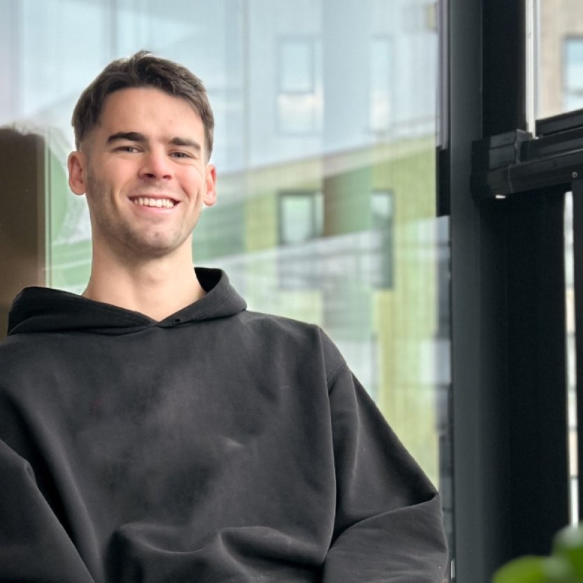 Mann i svart hettegenser smiler mens han sitter i en sofa ved et vindu.