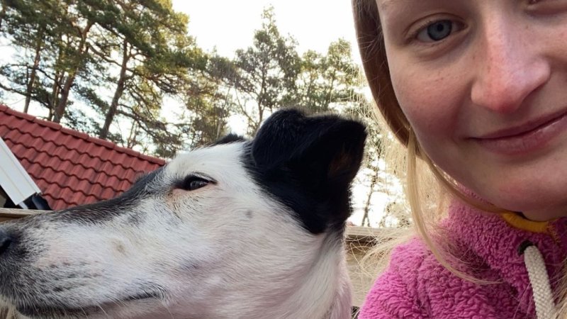 Kvinne i rosa genser smiler ved siden av en hund utendørs.