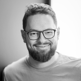 Mann med briller og skjegg smiler mot kamera i svart-hvitt bilde.