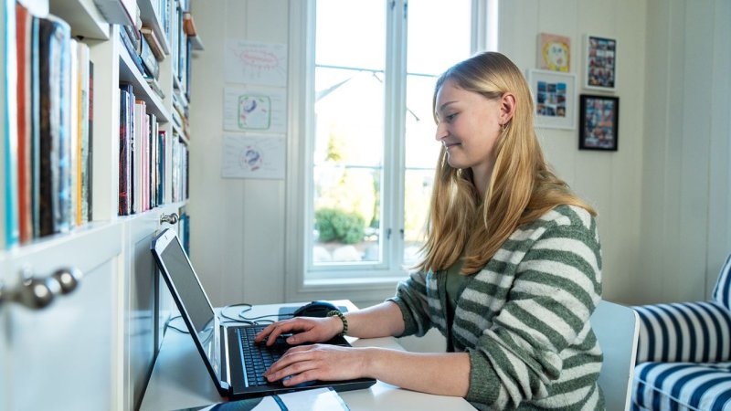 Kvinne studerer ved skrivebord med laptop i et lyst rom.