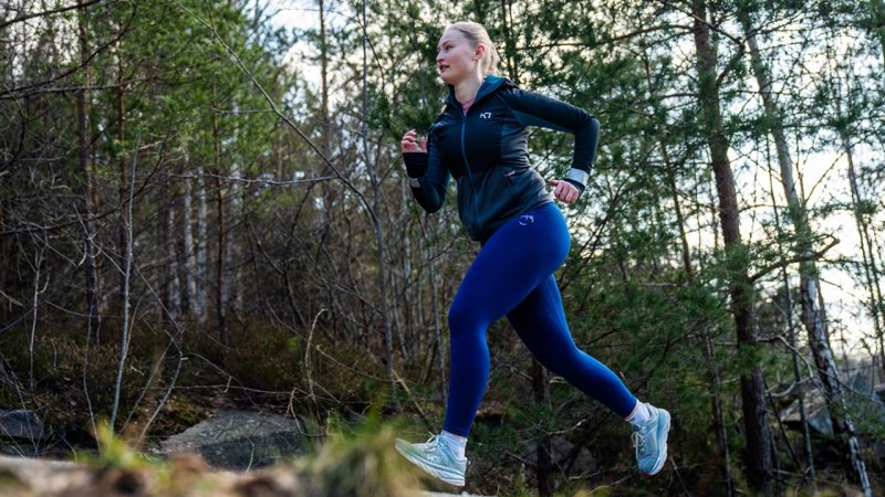 Kvinne jogger i skogen, iført treningsklær, omgitt av trær og natur.