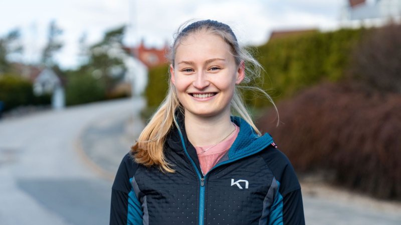 Ung kvinne smiler utendørs i sportsjakke på en solrik dag.