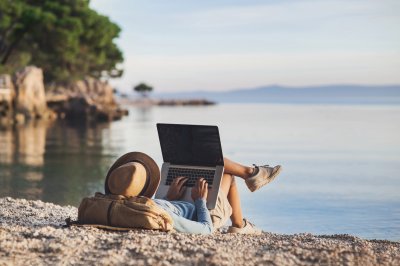 Person som studerer med laptop på strand, ryggsekk ved siden av.