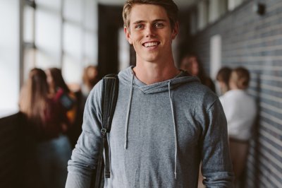 Ung mann i grå hettegenser smiler i en skolekorridor.