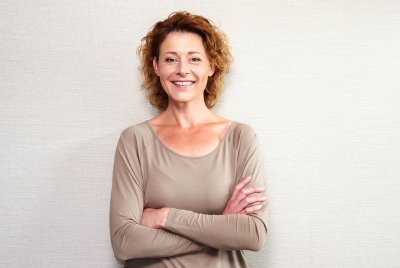 Kvinne med krøllete hår smiler og står med armene i kors foran en lys vegg.