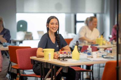 Kvinne smiler mens hun sitter ved et bord med medisinsk utstyr i et klasserom.
