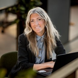 Kvinne med langt hår smiler mens hun jobber på en bærbar datamaskin.