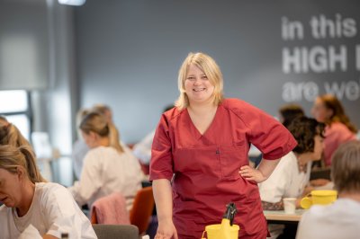 Kvinne i rød uniform smiler i et klasserom med andre studenter.