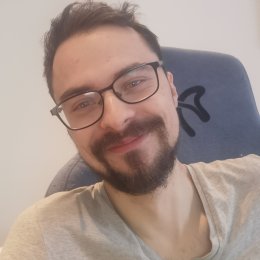 Mann med briller og skjegg smiler til kameraet, sitter i en stol.