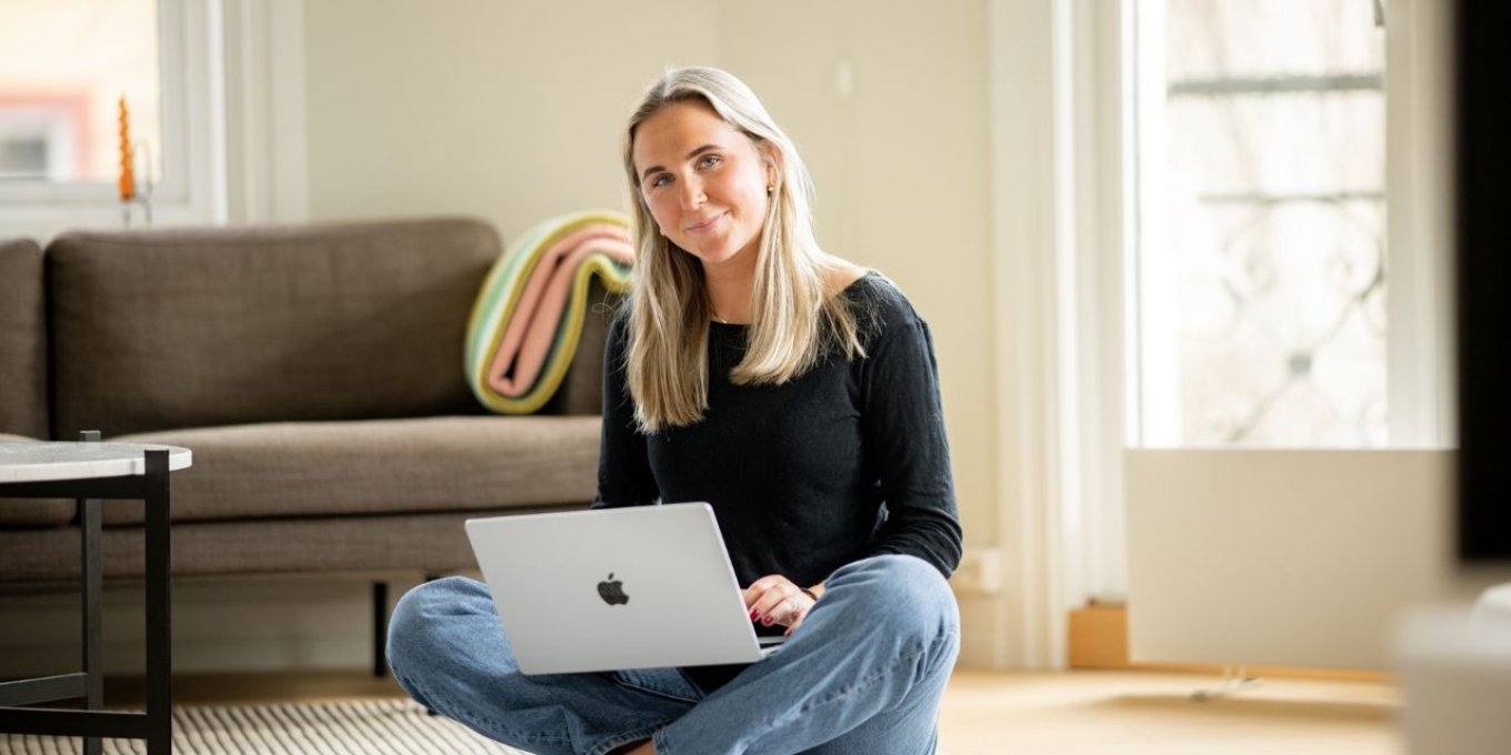 Kvinne sitter på gulvet med en laptop i fanget, smilende i et lyst rom.
