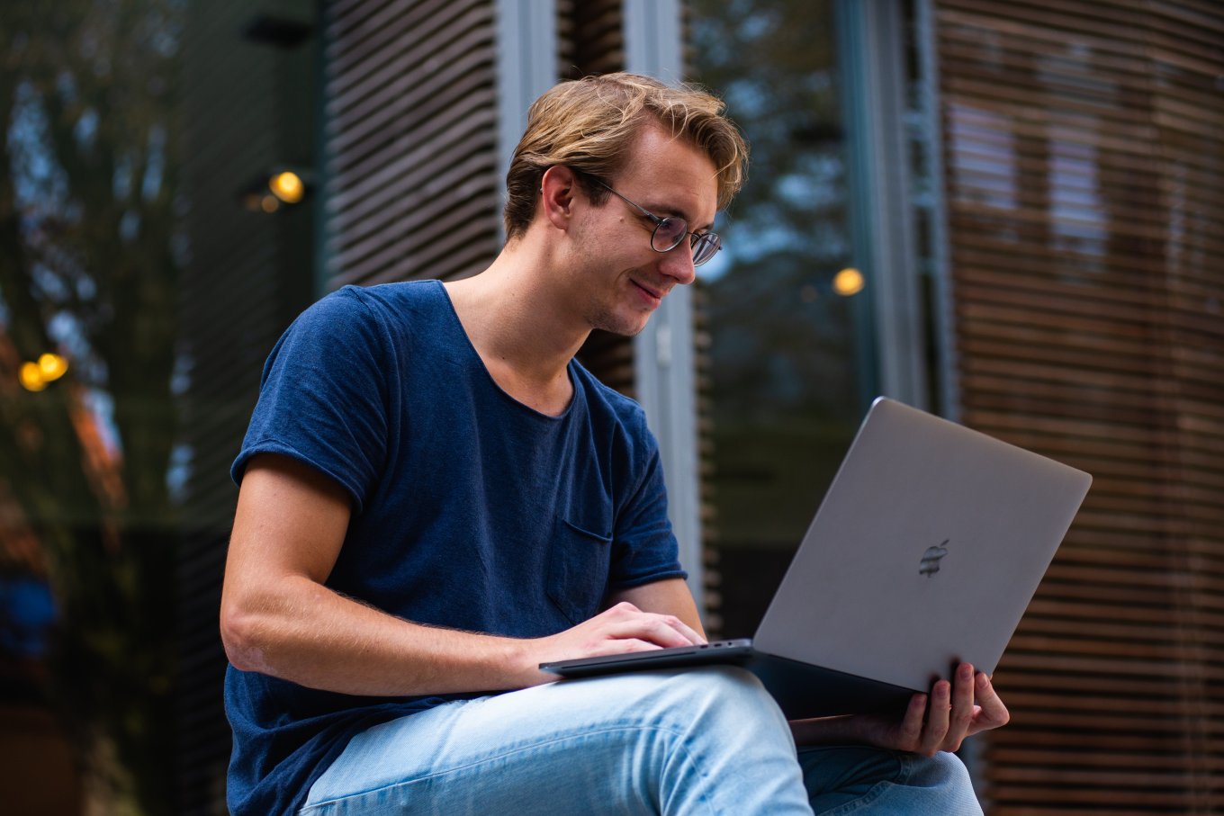 Mann studerer på laptop utendørs foran en bygning.