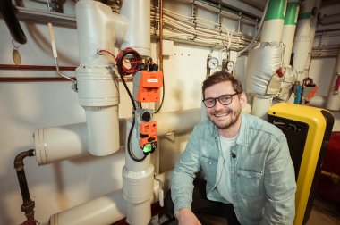 Mann smiler foran rør og teknisk utstyr i et maskinrom.