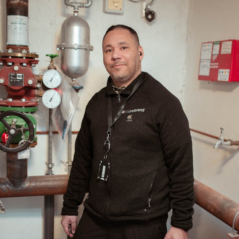 Person i arbeidsklær står foran rør og måleinstrumenter i teknisk rom.
