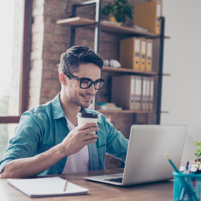 Mann med briller jobber på laptop og drikker kaffe ved skrivebord.